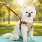 Un adorable petit chien blanc de type Spitz portant un harnais beige et marron avec un motif d'ours, et une laisse assortie, assis joyeusement dans un cadre extérieur verdoyant. L'image met en avant le "Confort & Style" du harnais.