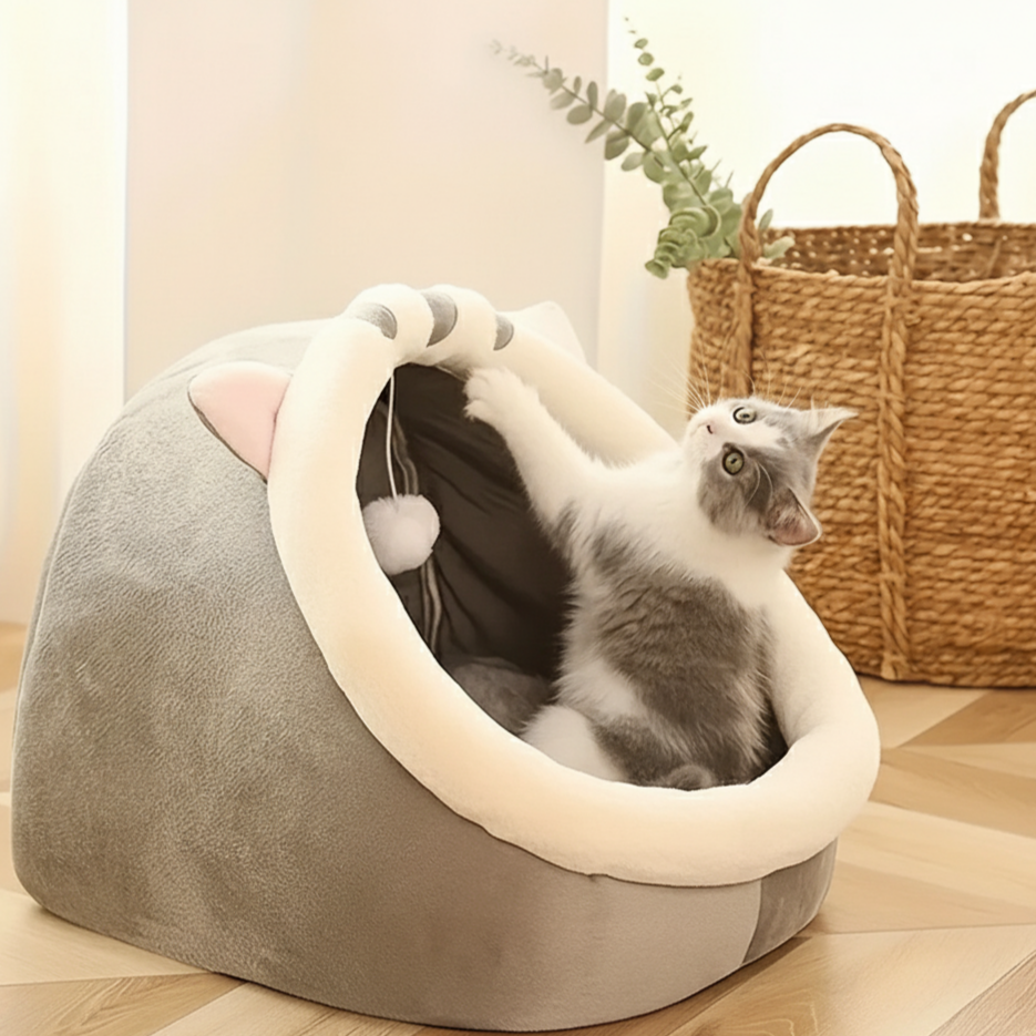 Un chat gris et blanc espiègle dans un lit douillet en forme de maisonnette, avec une balle suspendue, regardant vers le haut, capturant un moment de jeu confortable.